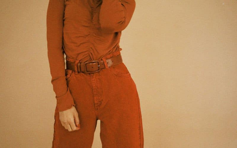 Femme portant une t-shirt manche longue orange brique et un pantalon orange brique lui aussi et une ceinture en cuir marron foncé sur un fond beige foncé. On ne voit ni sa tête, ni ses pieds.